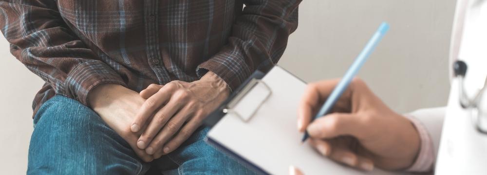 Doctor taking notes while man sits on doctors bench with his hands in his lap