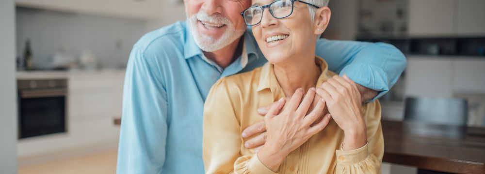 Older man hugging older woman wife with one arm in kitchen